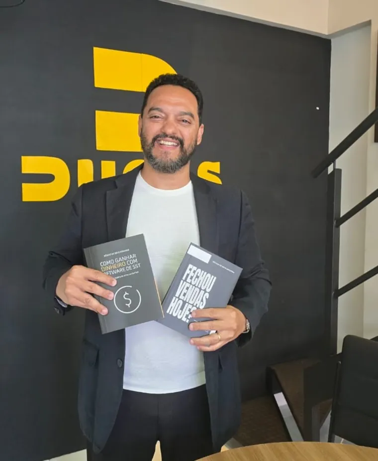 Sérgio Miranda segurando os livros “Como Ganhar Dinheiro com Software de SST” e “Fechou Vendas Hoje?” em frente ao painel da DUOSS.